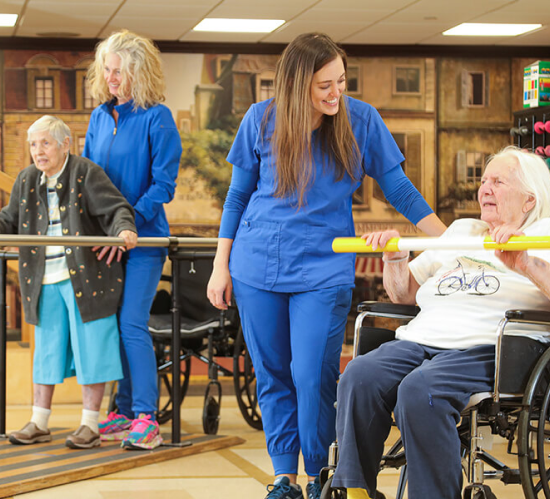 Caregiver in blue scrubs assisting a seated resident in a wheelchair, with two other seniors in the background