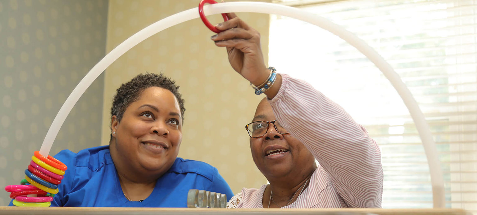 Two people smiling as one threads a red ring onto a white arch in a bright room