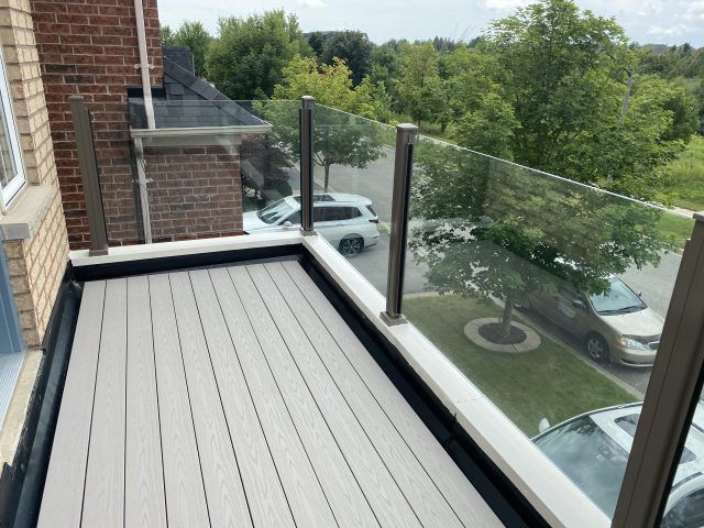 A balcony with a glass railing and a brick building in the background by Premier Decks & Awnings