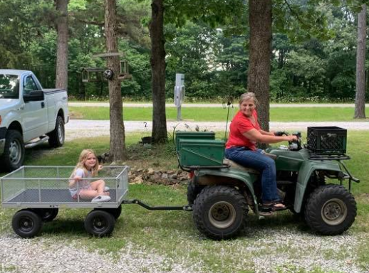 Person on green ATV towing child in a small wagon in a wooded yard