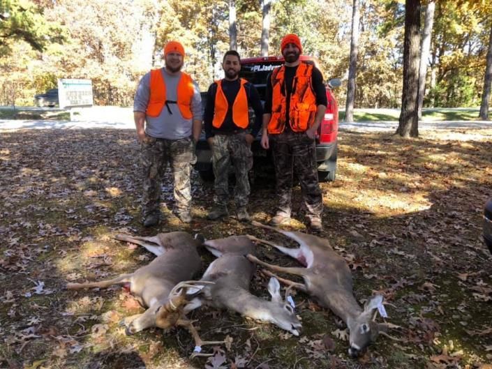 Three hunters in orange vests stand behind several downed deer in a wooded area.