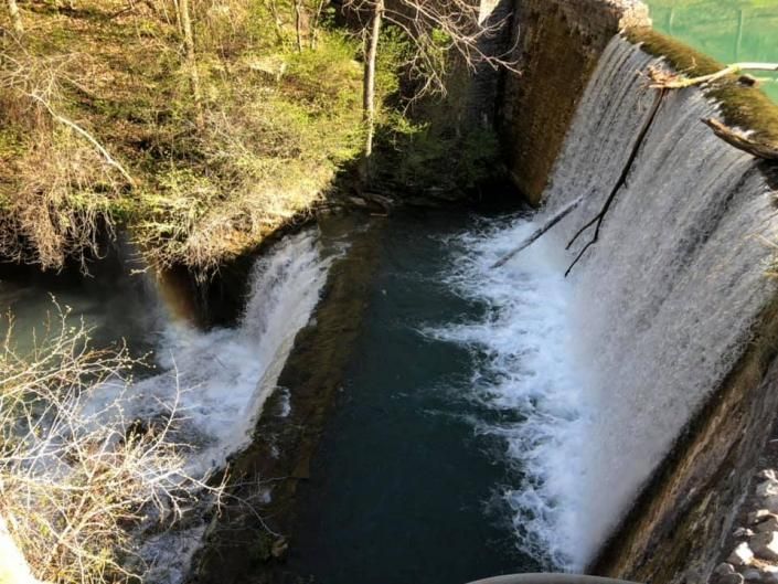 Water cascading over a curved dam into a dark river below, surrounded by rocky banks and dry grass.