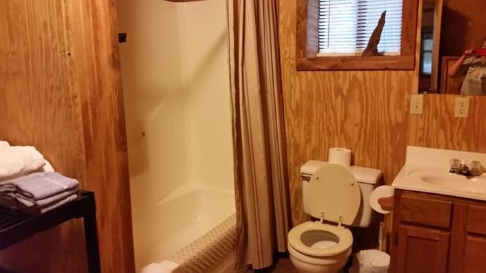 Compact wood-paneled bathroom with a toilet, sink, shower, and window.