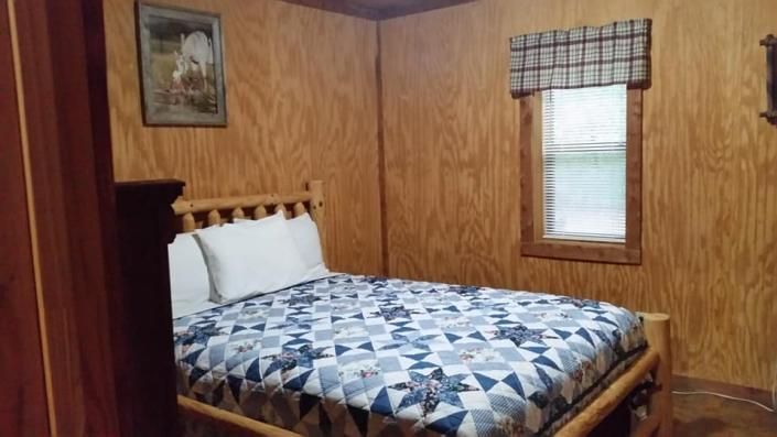 Cozy wood-paneled bedroom with a quilted bed, framed wall art, and a small window with plaid curtains
