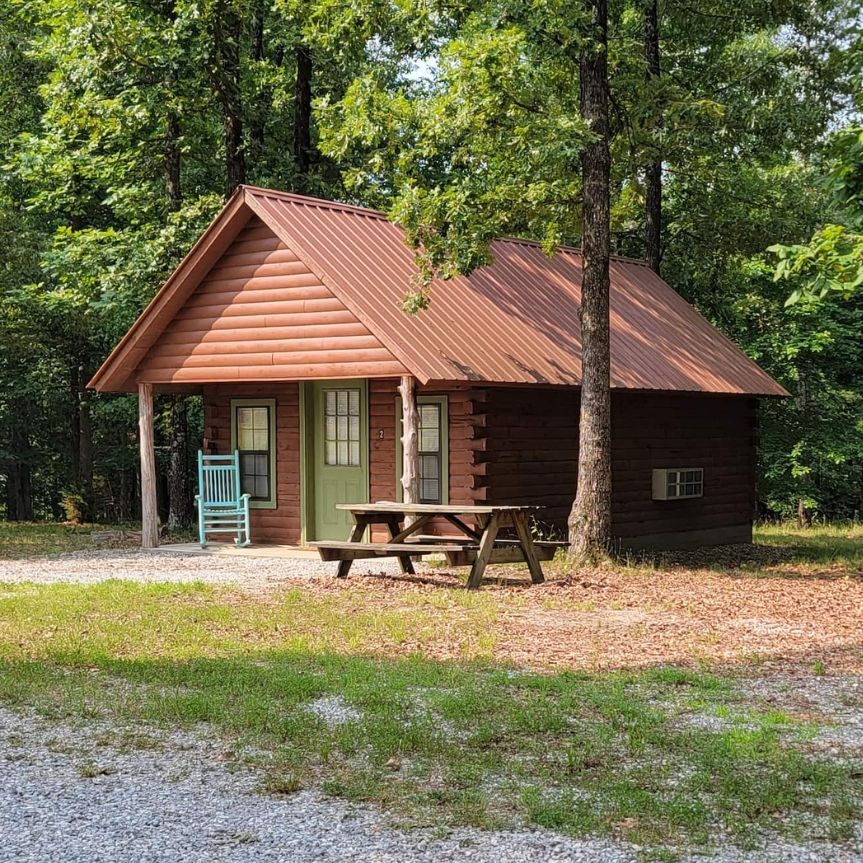Small rustic cabin with a red roof and porch, surrounded by trees and a picnic table in a wooded clearing