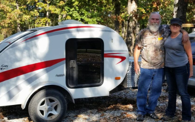 Man and woman standing beside a white teardrop camper with red stripes in a wooded area