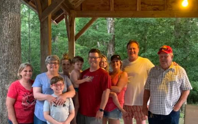 Group of people smiling under a wooden pavilion at dusk, with trees in the background.
