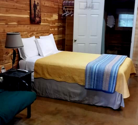 Cozy rustic bedroom with a bed, yellow and blue bedding, wooden walls, and a lamp beside a chair