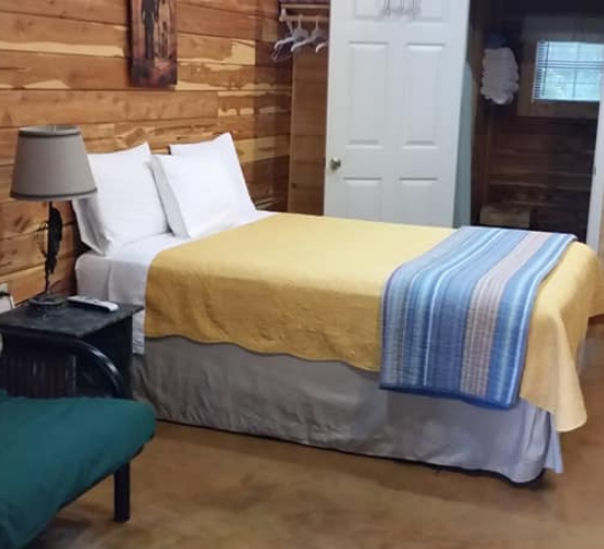 Rustic bedroom with a made bed, white pillows, yellow blanket, blue runner, and bedside lamp