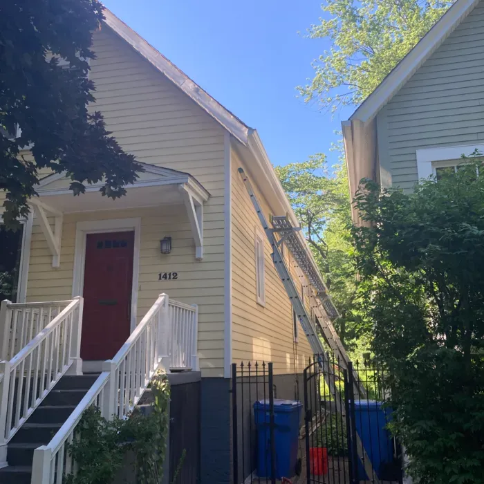 Yellow house with a ladder propped against the side, blue trash bins in front of a black fence.