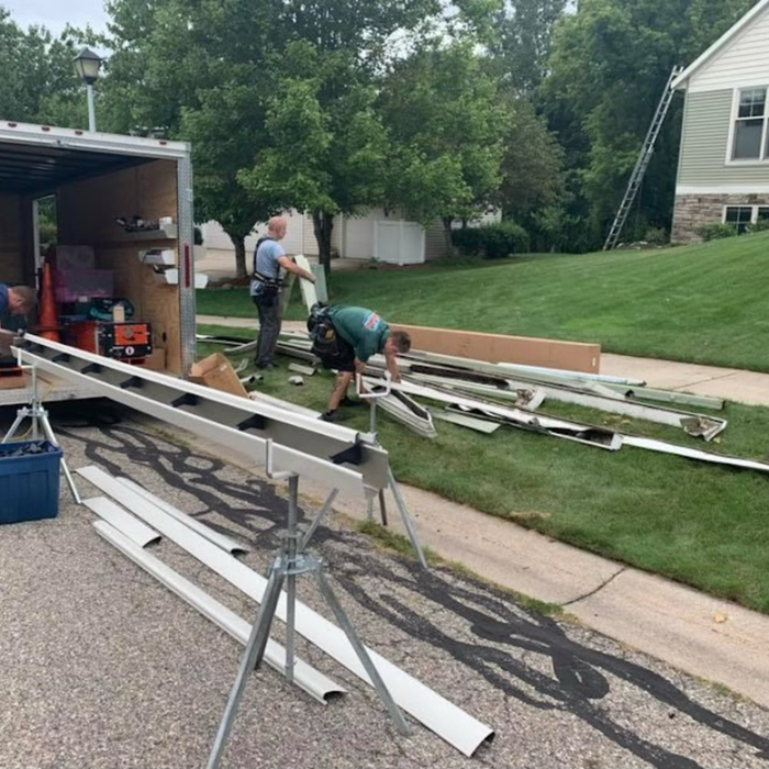 Workers unloading materials from a trailer on a residential driveway. Gutters on sawhorses.