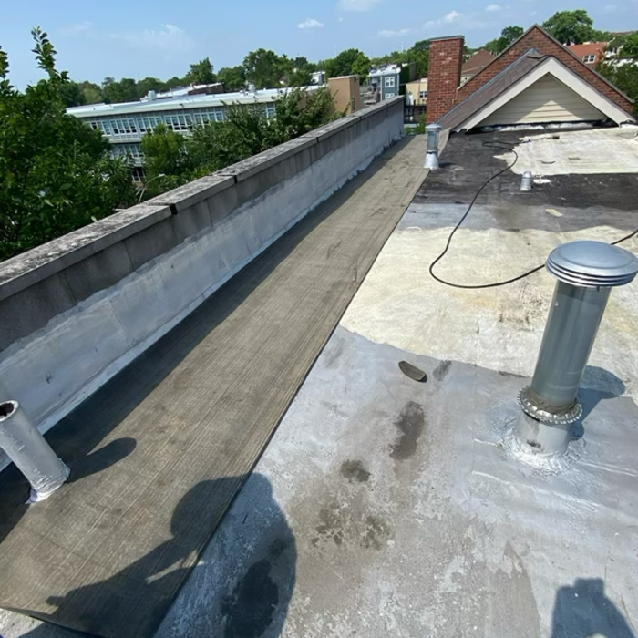 Flat rooftop with various ventilation pipes, a concrete barrier, and a walkway. Buildings and trees in the background.