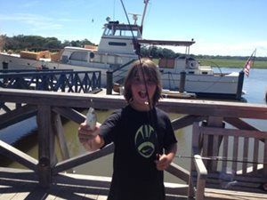 Child on a dock holding a fish, with boats and water in the background.