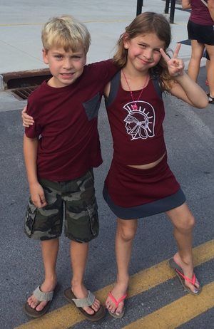 Two children standing arm-in-arm on a boardwalk, smiling and wearing maroon shirts and sandals.