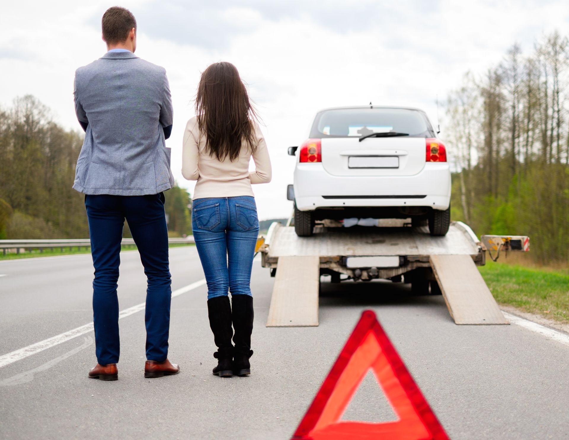 Broken-down white car on tow truck ramp with warning triangle on roadside