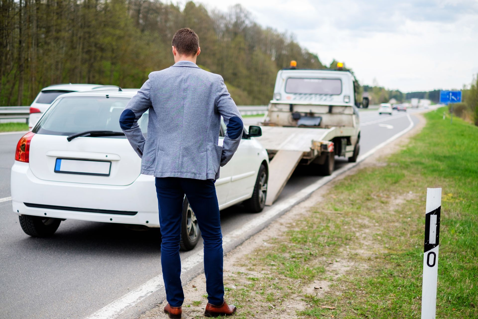 Man standing by a white car on a roadside, with a tow truck ahead on the shoulder
