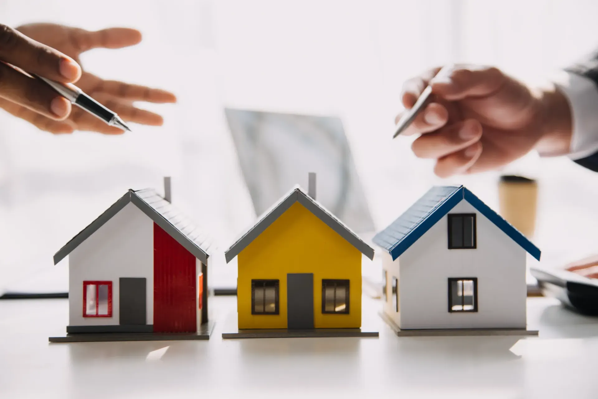 Three miniature houses with hands holding pens, discussing real estate.