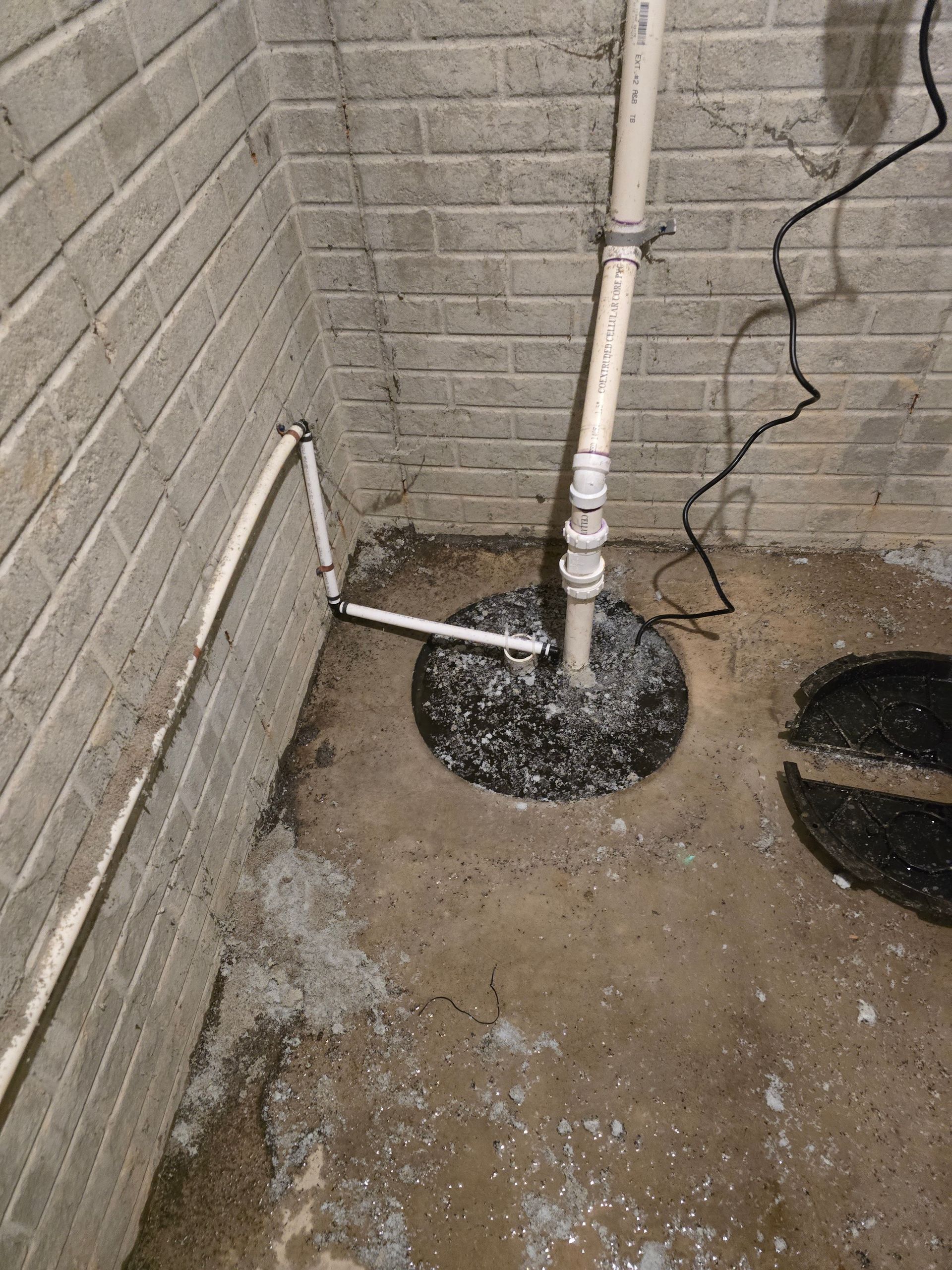Basement sump pump system: white pipes, black pump, gray concrete floor, and light brick walls.