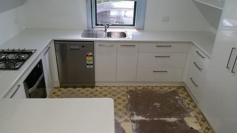 White kitchen with countertops, cabinets, appliances, and a table. Damaged floor and a window.
