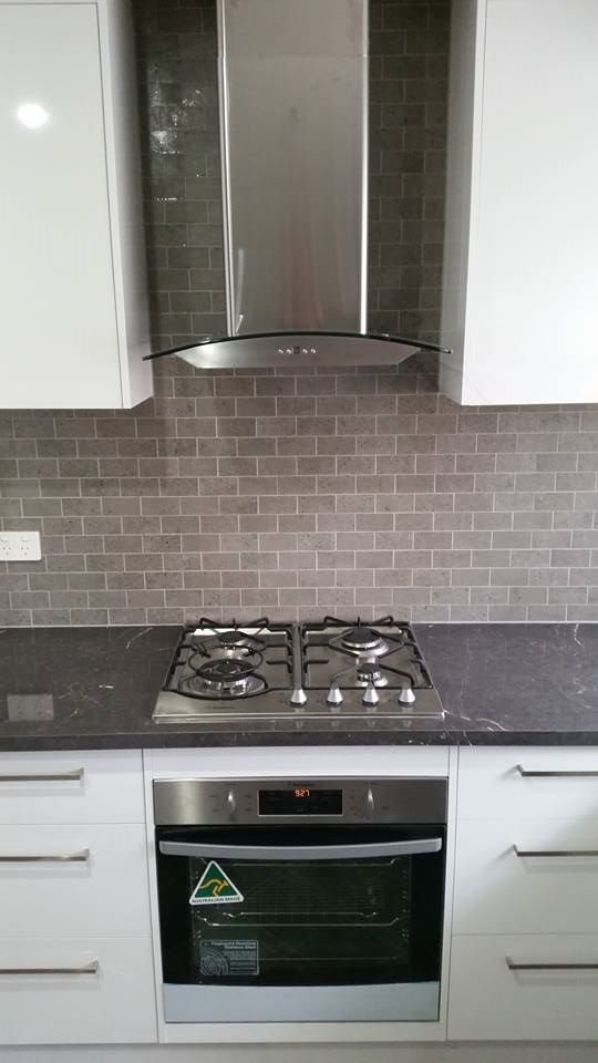 Modern kitchen with stove, oven, range hood, and white cabinets. Gray tile backsplash and black countertop.