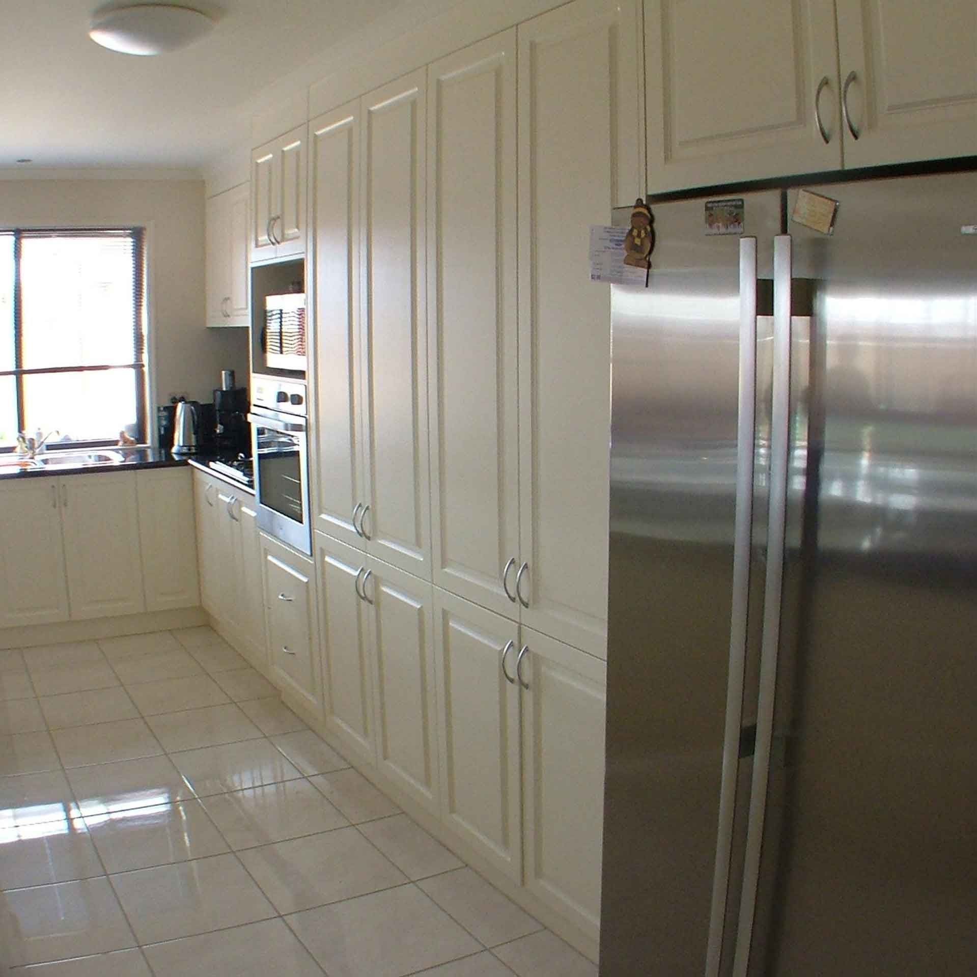 Cream-colored kitchen with tall cabinets, stainless steel refrigerator, and tiled floor. Window in the corner with black countertop.