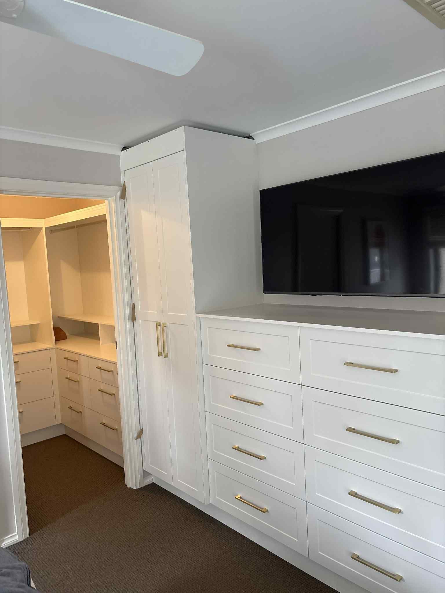 White custom closet unit with drawers and a TV above, next to a walk-in closet.