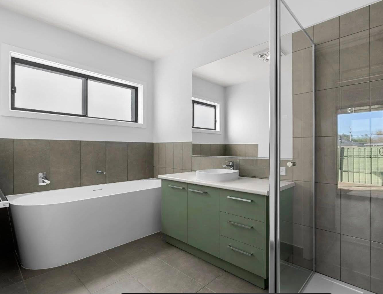 Modern bathroom with a freestanding white bathtub, green vanity, and a glass shower. Light gray walls and tiled accents are visible.