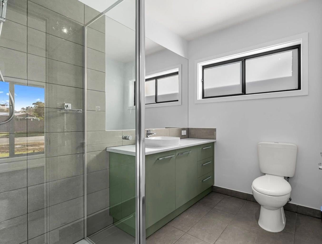 Modern bathroom with a glass shower, sage green vanity, white toilet, and a window overlooking a neighborhood.