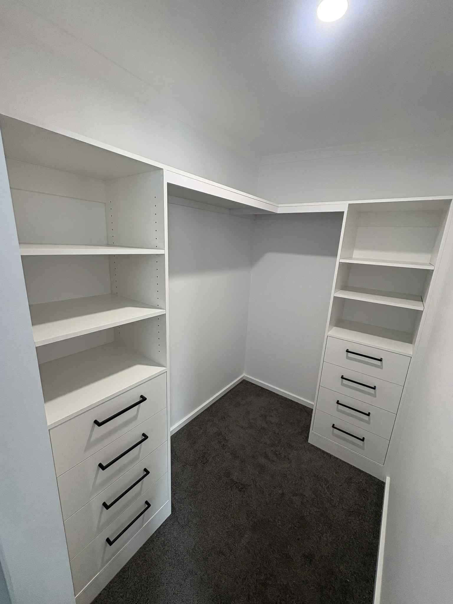 Empty white closet with built-in shelving and drawers, dark carpeted floor, and bright overhead light.