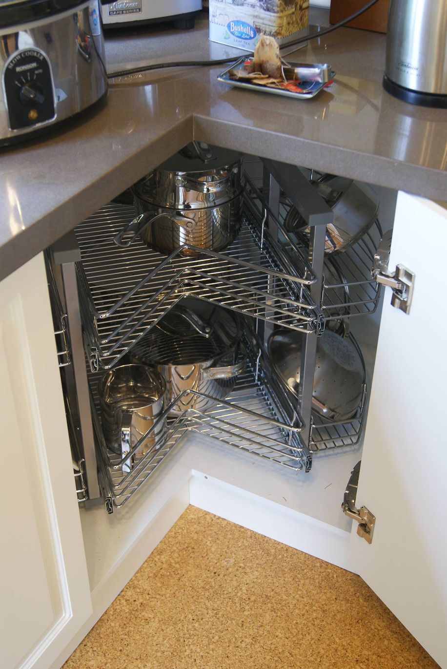 Corner kitchen cabinet with pull-out shelving holding pots and pans. Off-white doors, cork flooring, gray countertop.