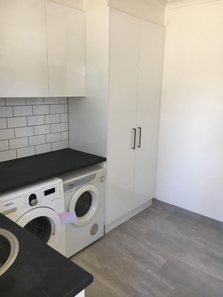 Laundry room with white cabinets, black countertop, washing machine, and white brick-like backsplash. Gray floor and a tall cabinet are also present.