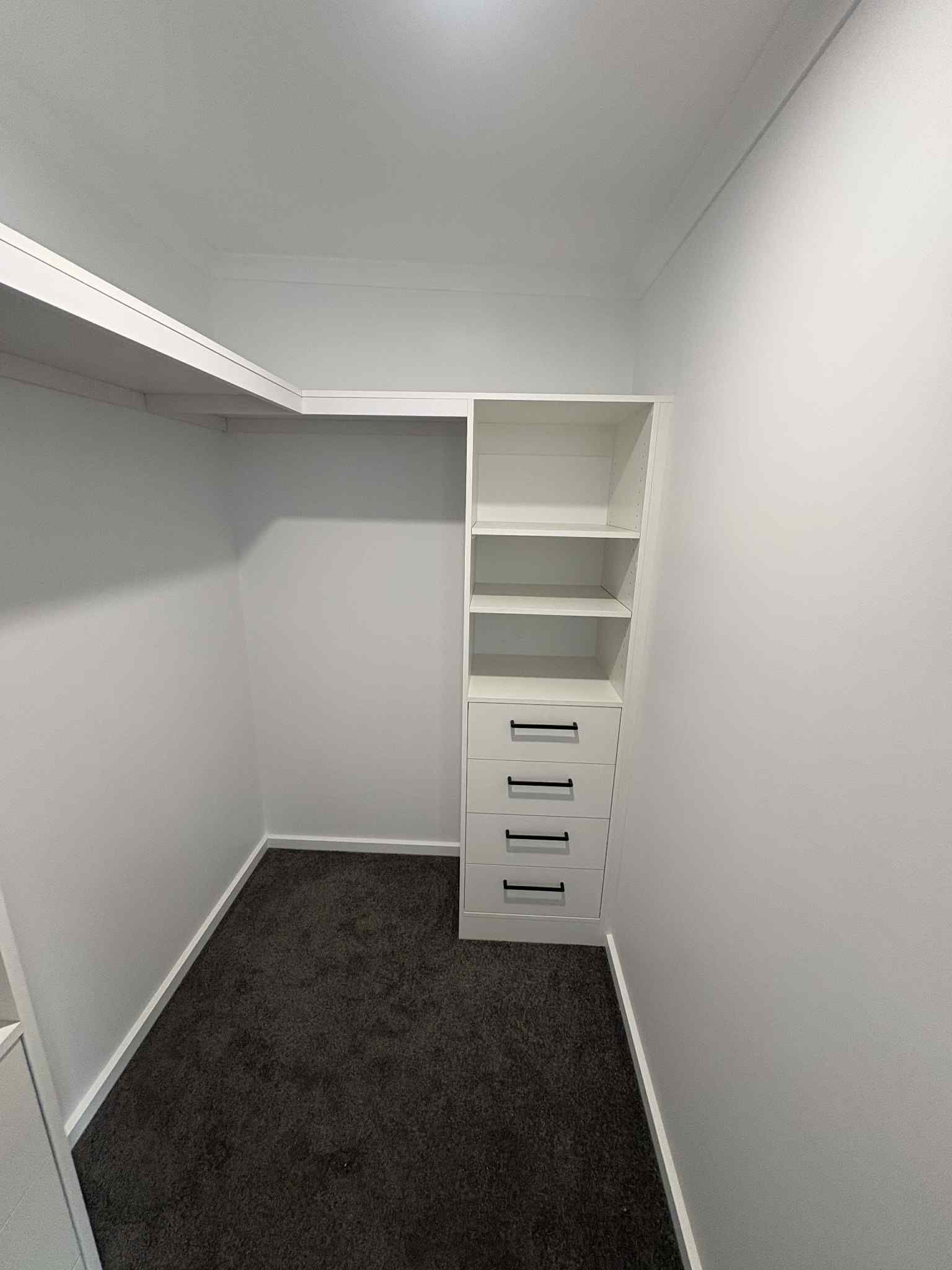 Empty walk-in closet with white shelves and drawers, dark gray carpet, white walls.