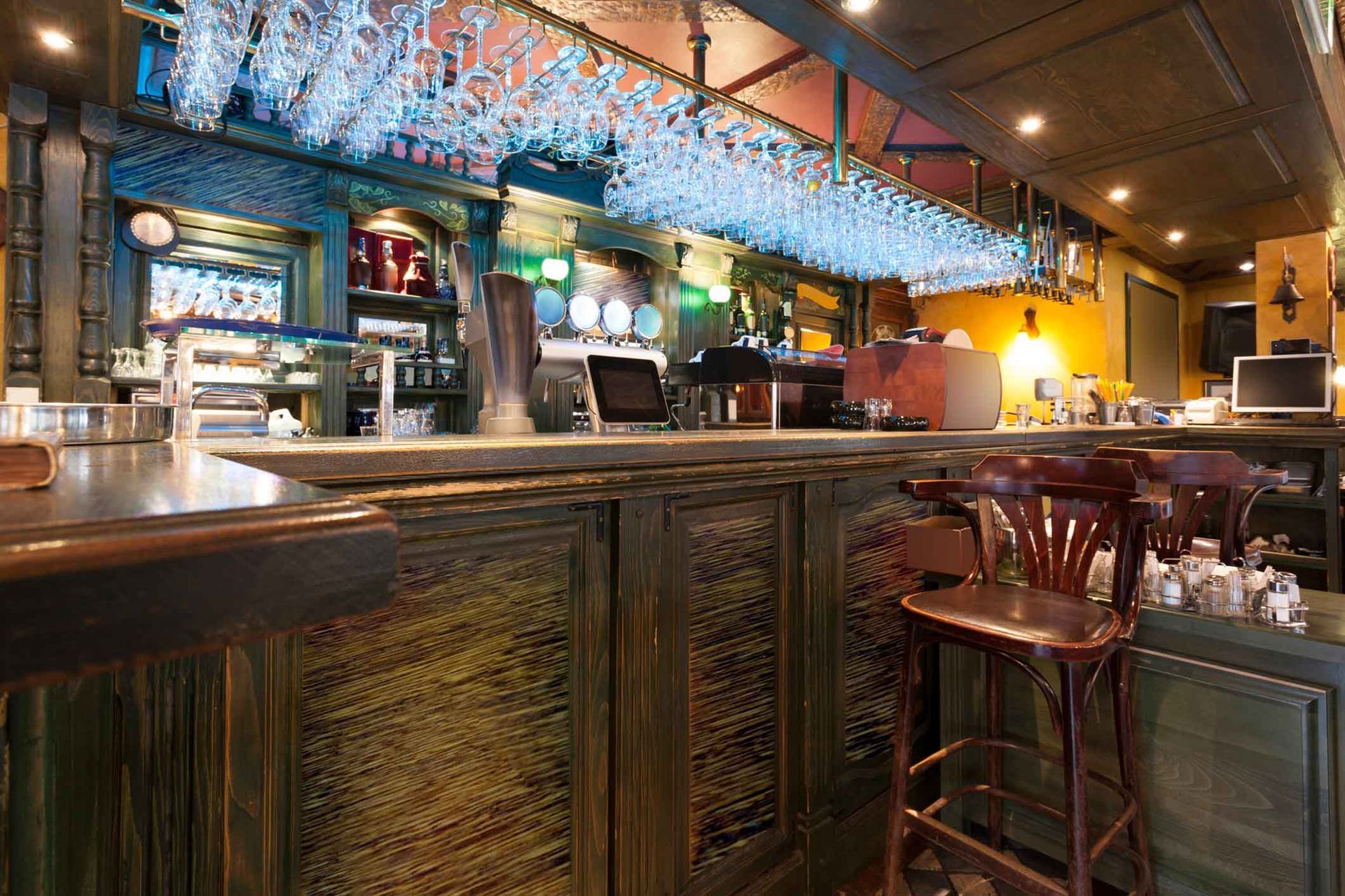 Wooden bar in a dimly lit pub, with stools, hanging glasses, and bottles.