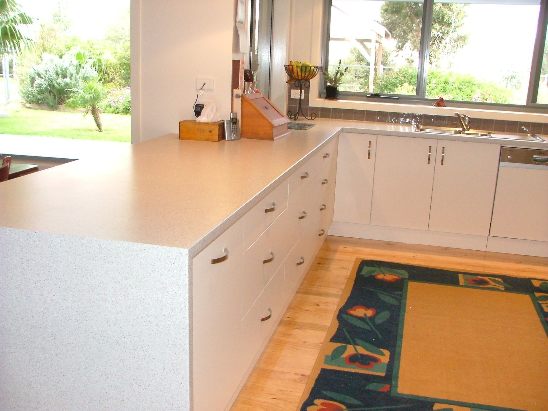 Kitchen with white countertops, cabinets, and a rug on wooden floors. Window view of a yard.