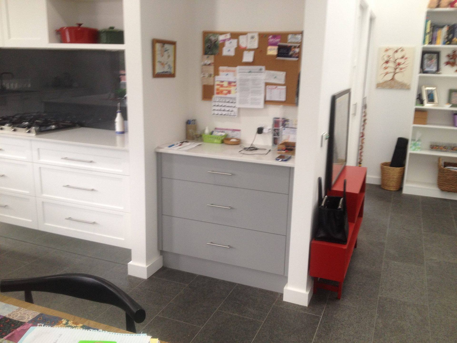 Kitchen area with white cabinets, gray drawers, corkboard, red shelf, and black chair.