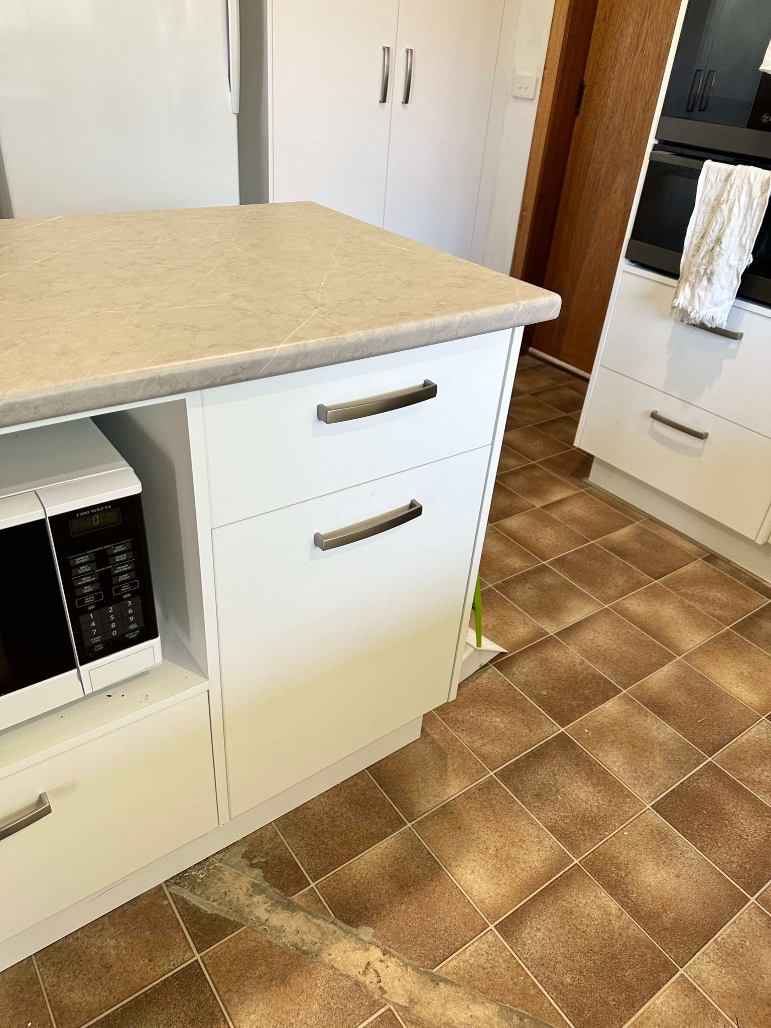 White kitchen cabinet with microwave and drawers, tan countertop, brown tiled floor.