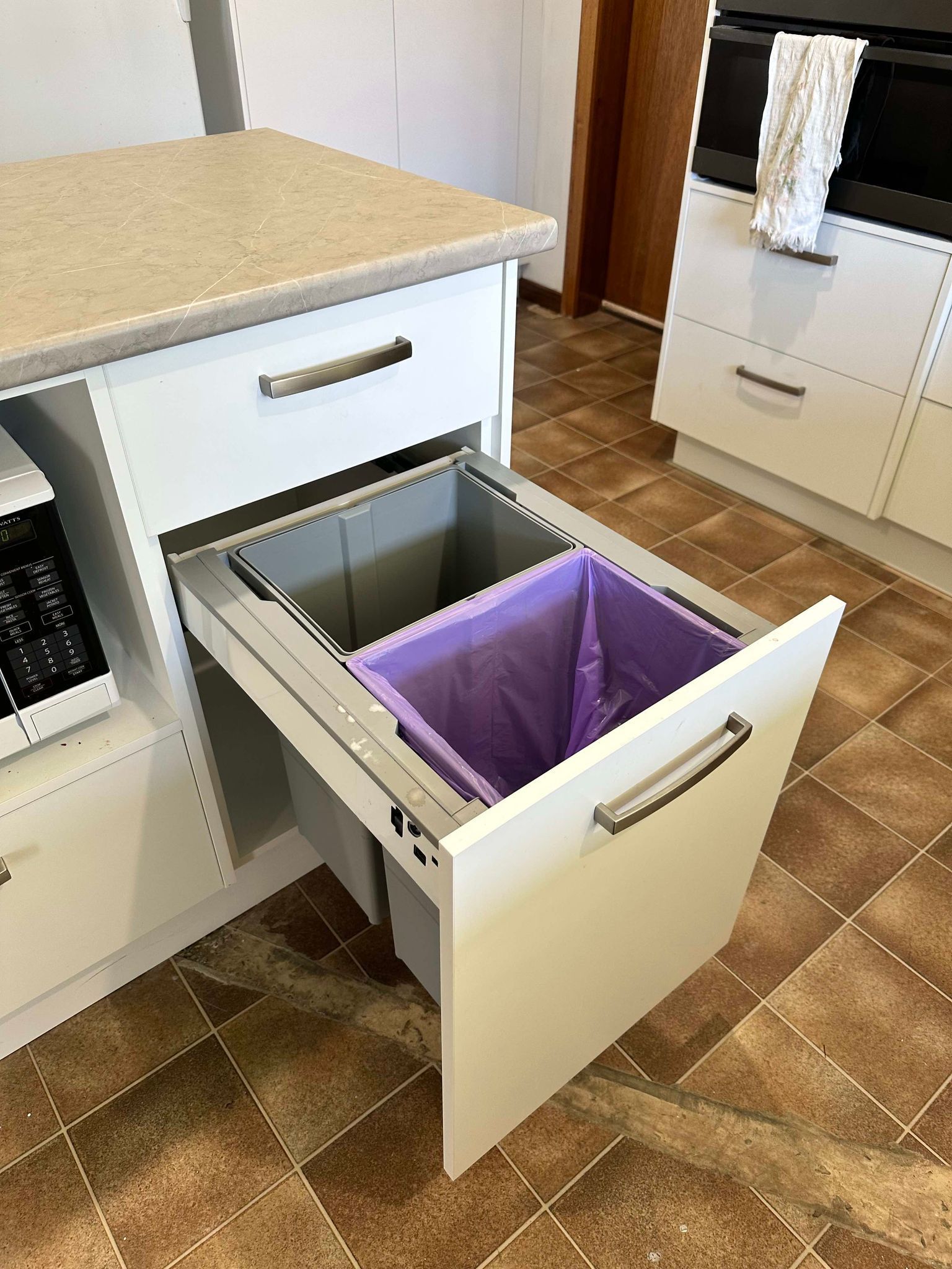 White kitchen cabinet with pull-out trash bins, one with a purple trash bag. Brown floor.