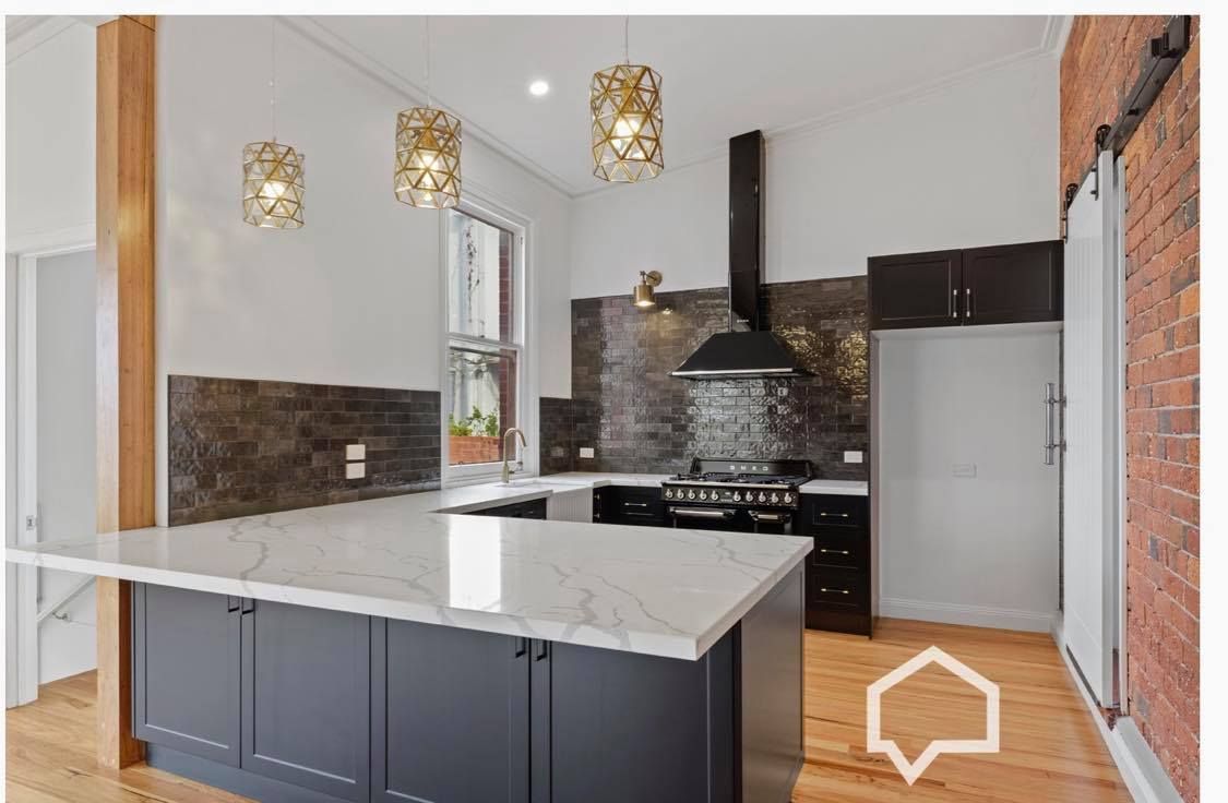 Modern kitchen with marble countertop island, dark cabinets, gold light fixtures, and brick wall.