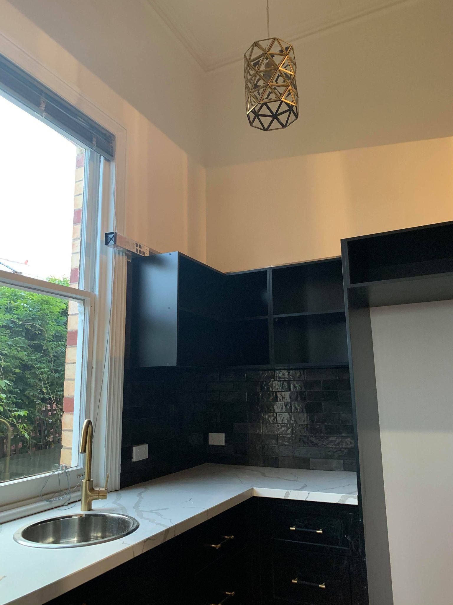 A corner kitchen with black cabinetry, tiled backsplash, and a gold sink by a window.