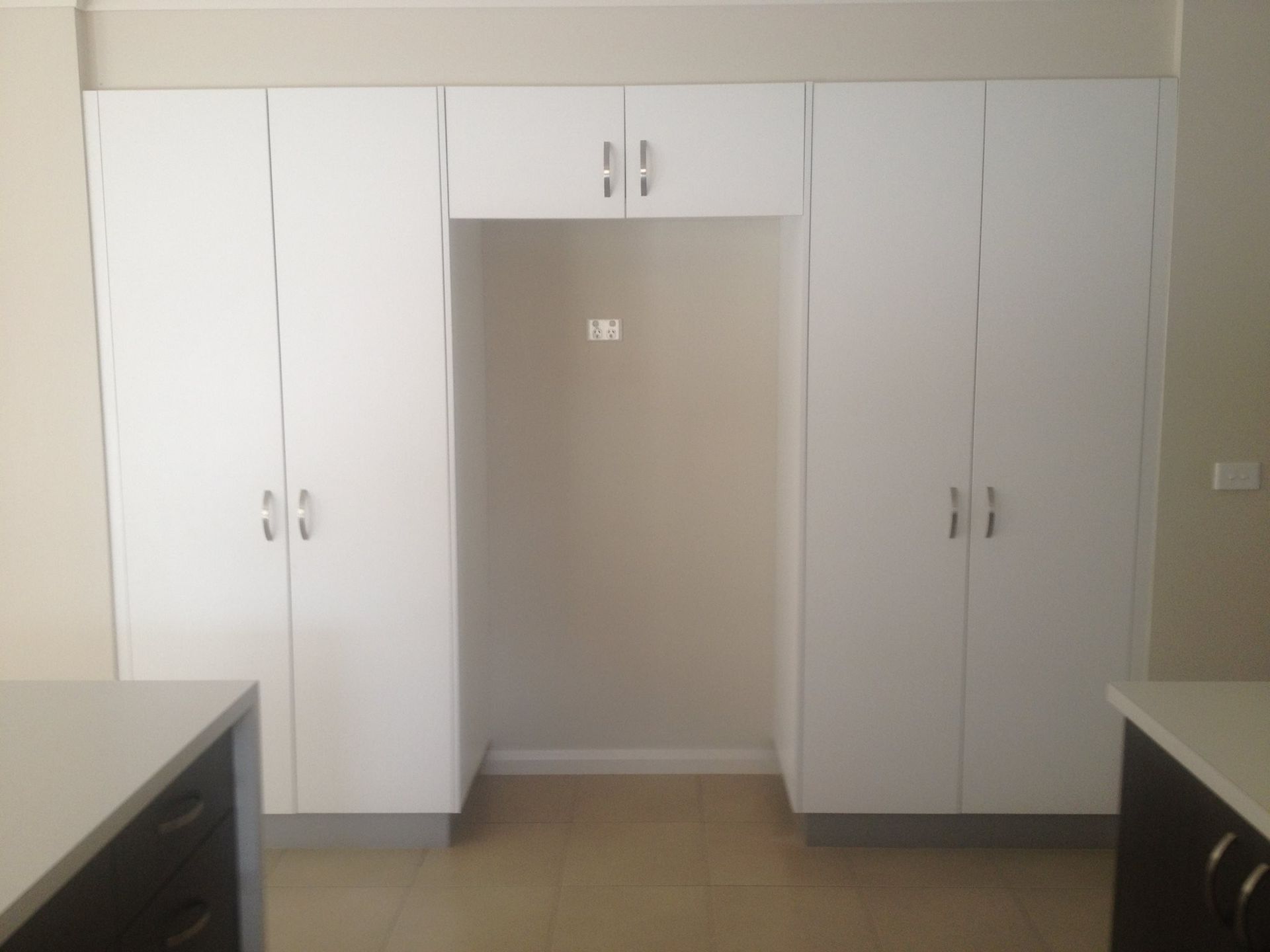 White kitchen cabinets surround a recessed area for a refrigerator, beige wall.