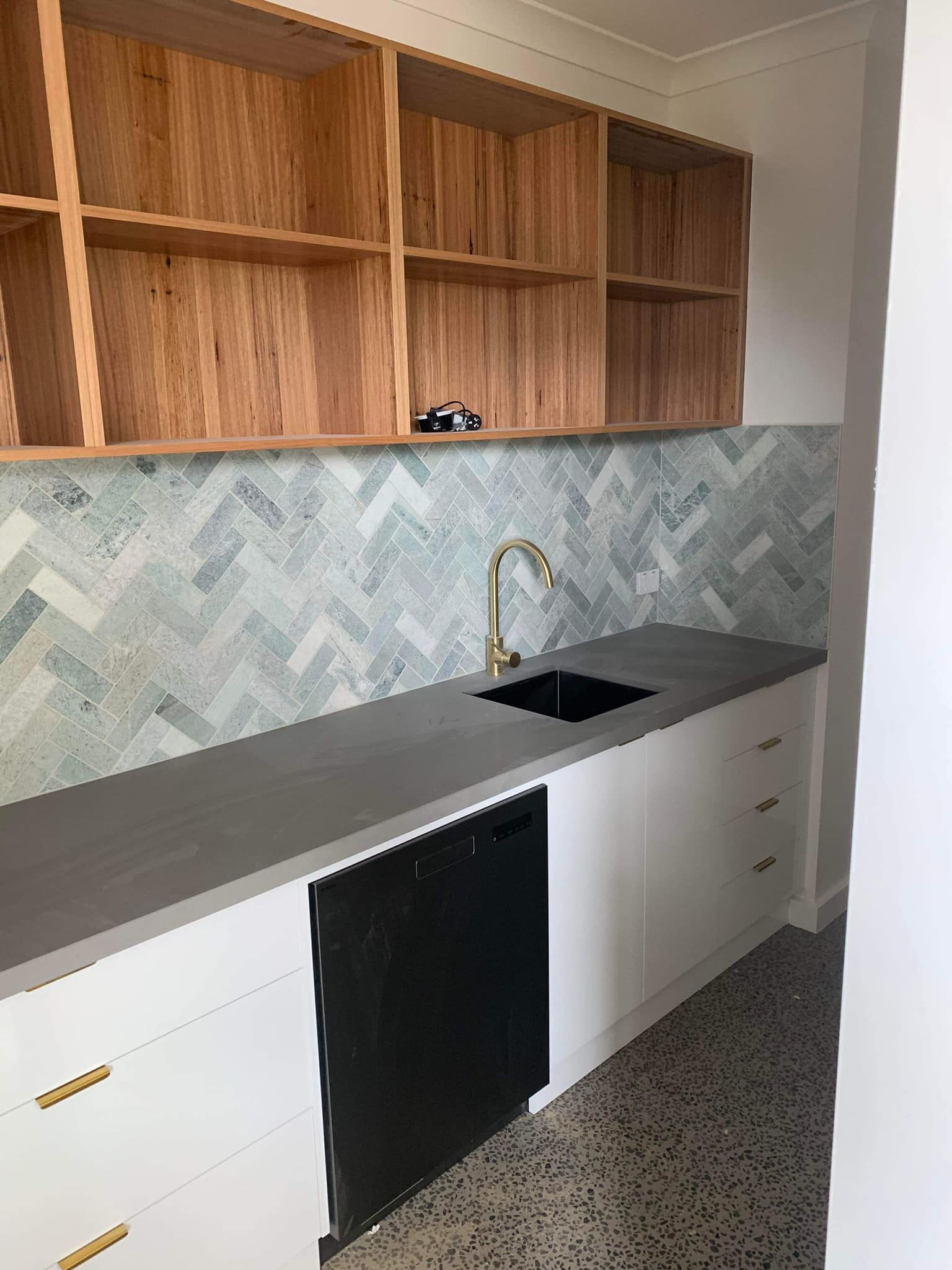 A modern laundry room with white cabinets, gray countertop, black appliances, and patterned backsplash. Wooden shelves are above.
