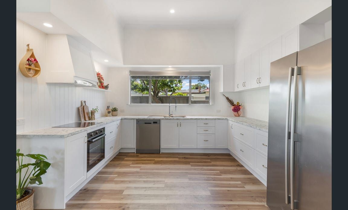 White kitchen with wooden floors, stainless steel appliances, and a large window overlooking trees.