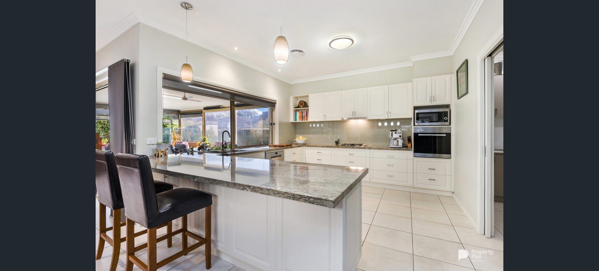 A bright kitchen with white cabinets, a granite island, and stools. Large windows look out to a garden.