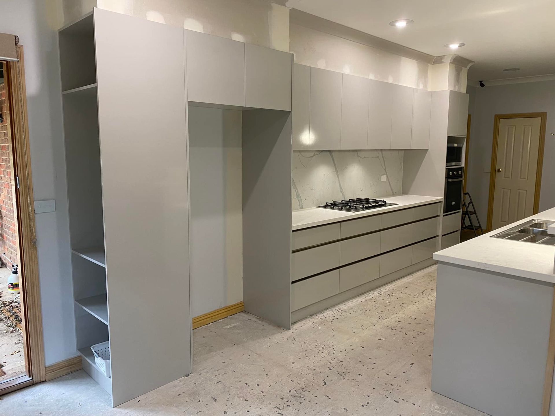 Modern kitchen under construction with gray cabinets, white countertops, and open refrigerator space.