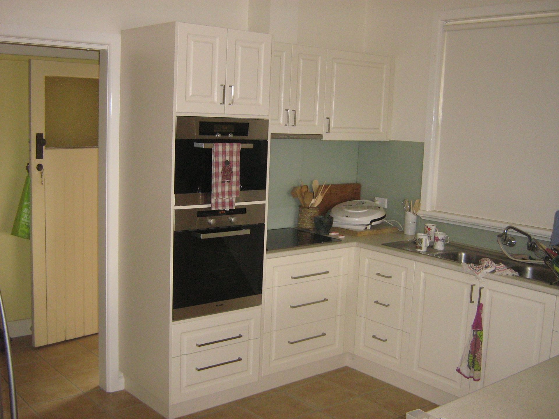 White kitchen cabinets with built-in oven and drawers. Corner sink area with light green backsplash.