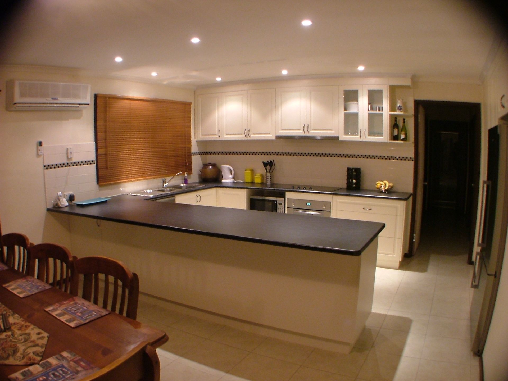Kitchen with cream cabinets, black countertop, and brown blind over the window, next to a dining area.