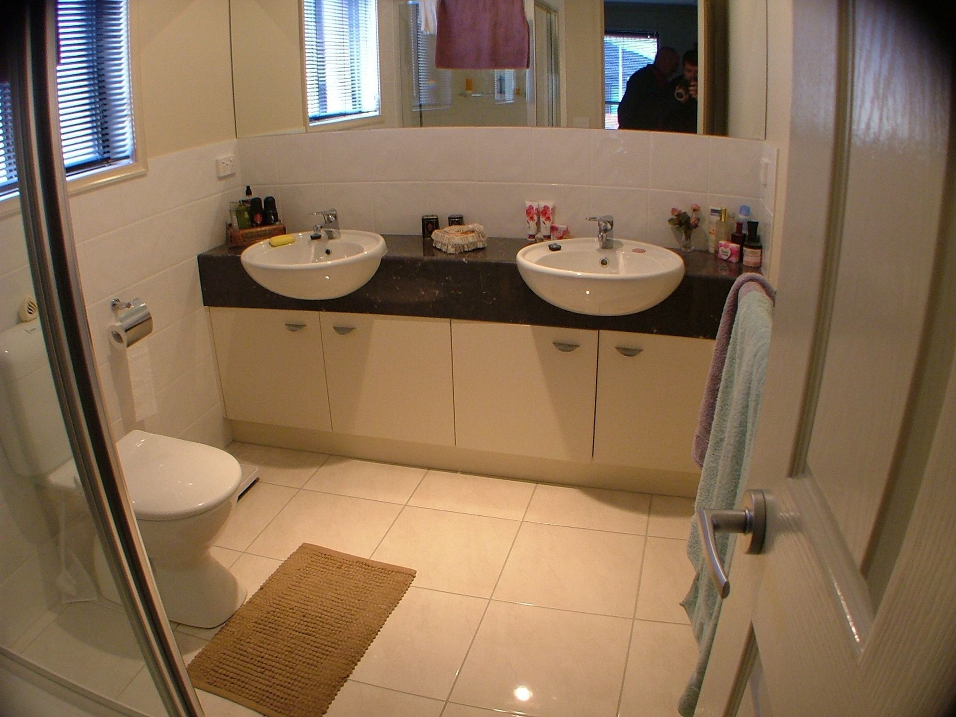 Bathroom with two sinks on a cabinet, toilet, shower, and tan rug on tile floor.