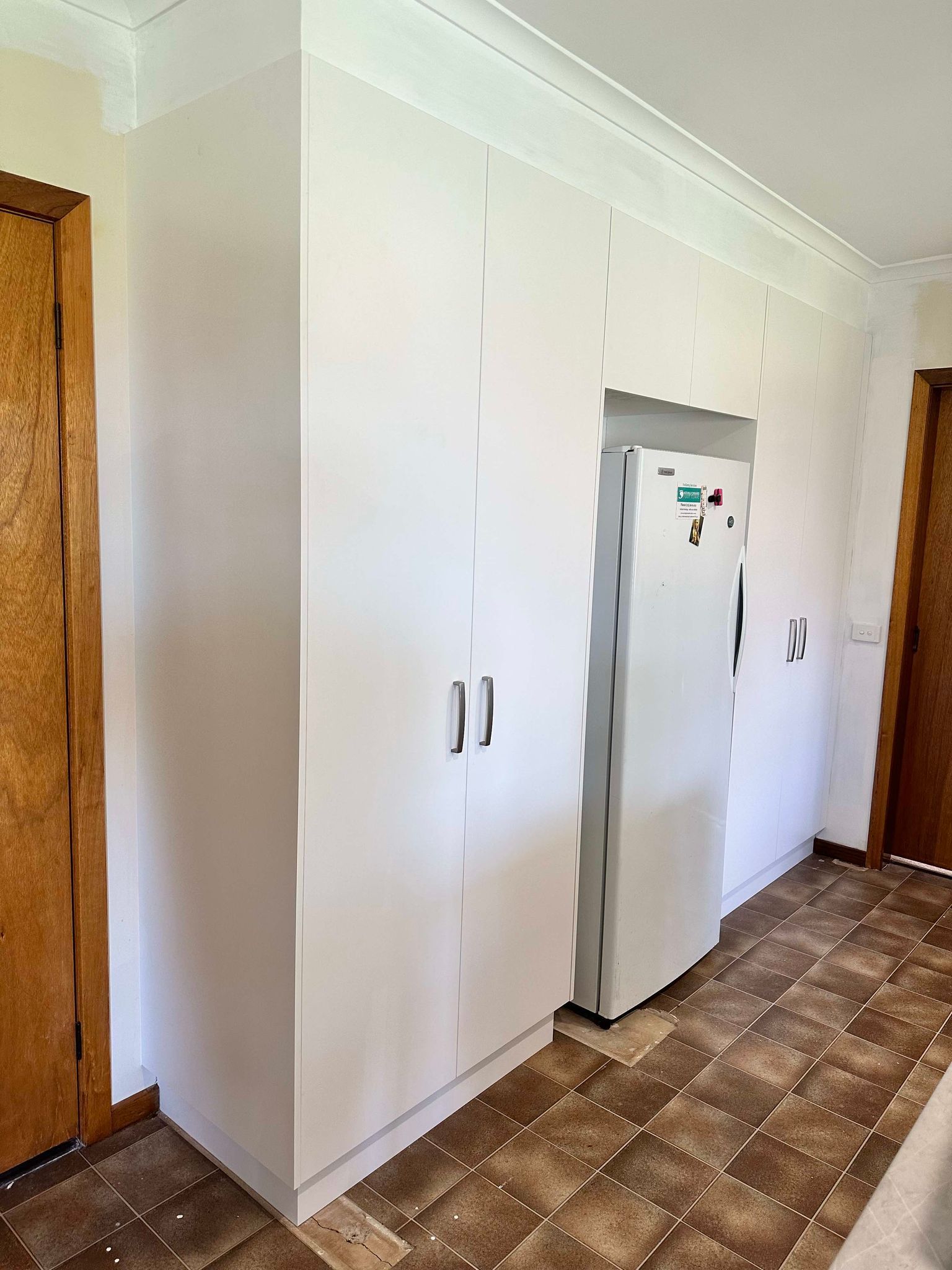 White built-in kitchen cabinets and refrigerator on a patterned floor. Two wooden doors are visible.