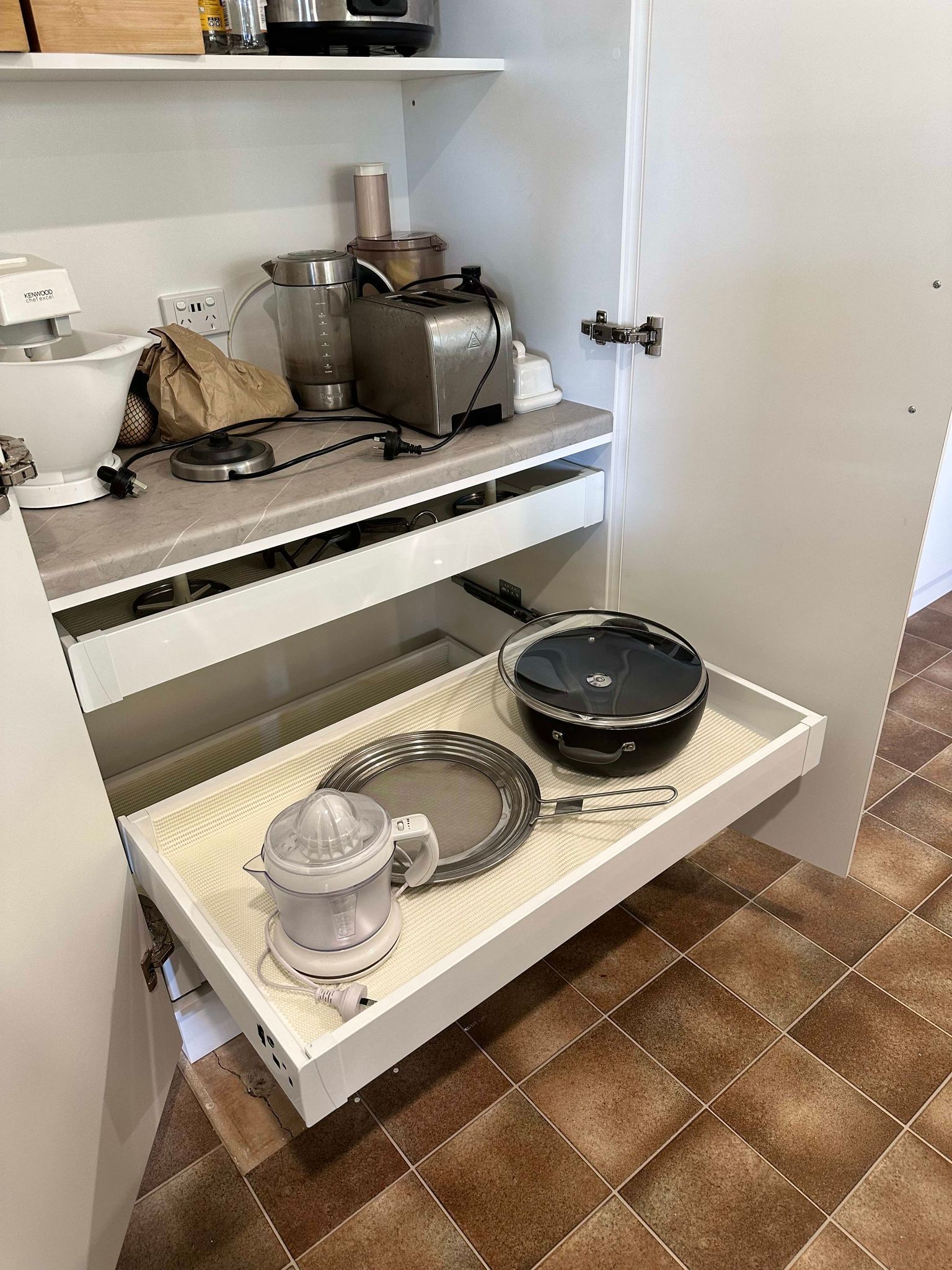 White cabinet with pull-out drawer holding kitchen items: juicer, plates, and pot. Interior countertop with small appliances.