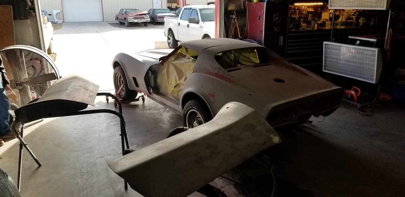 Classic sports car body in a garage, partially assembled with large detached panels and tools nearby.